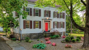 A two-story house with a red door, stone foundation, and a front garden with various plants and flowers. - Relax in Style at Historic Modern on Main With Modern Amenities & Wooded Views (Stone Ridge)