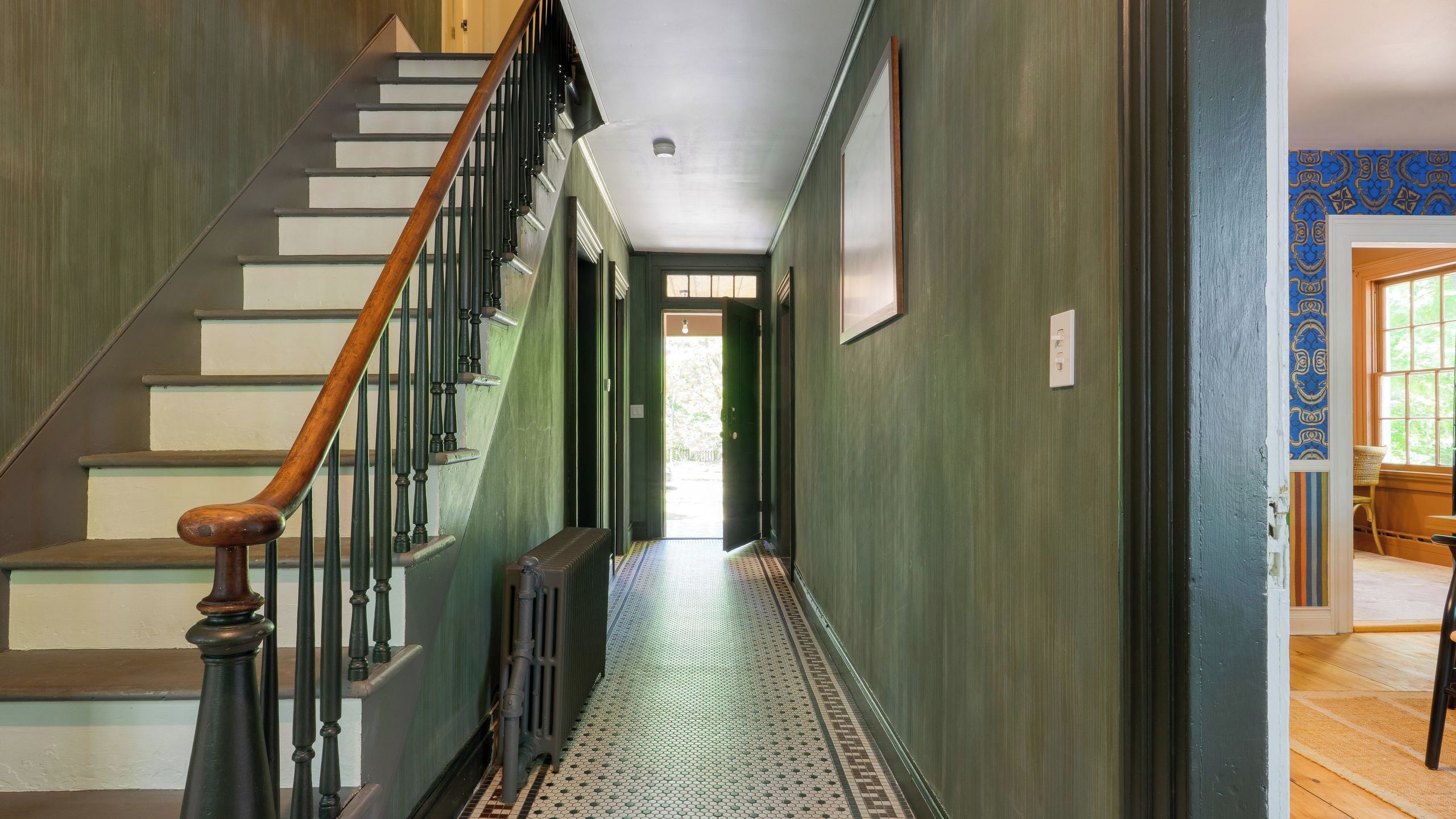 A hallway with a wooden staircase, a radiator, patterned floor tiles, and a door leading to a room with patterned wallpaper.