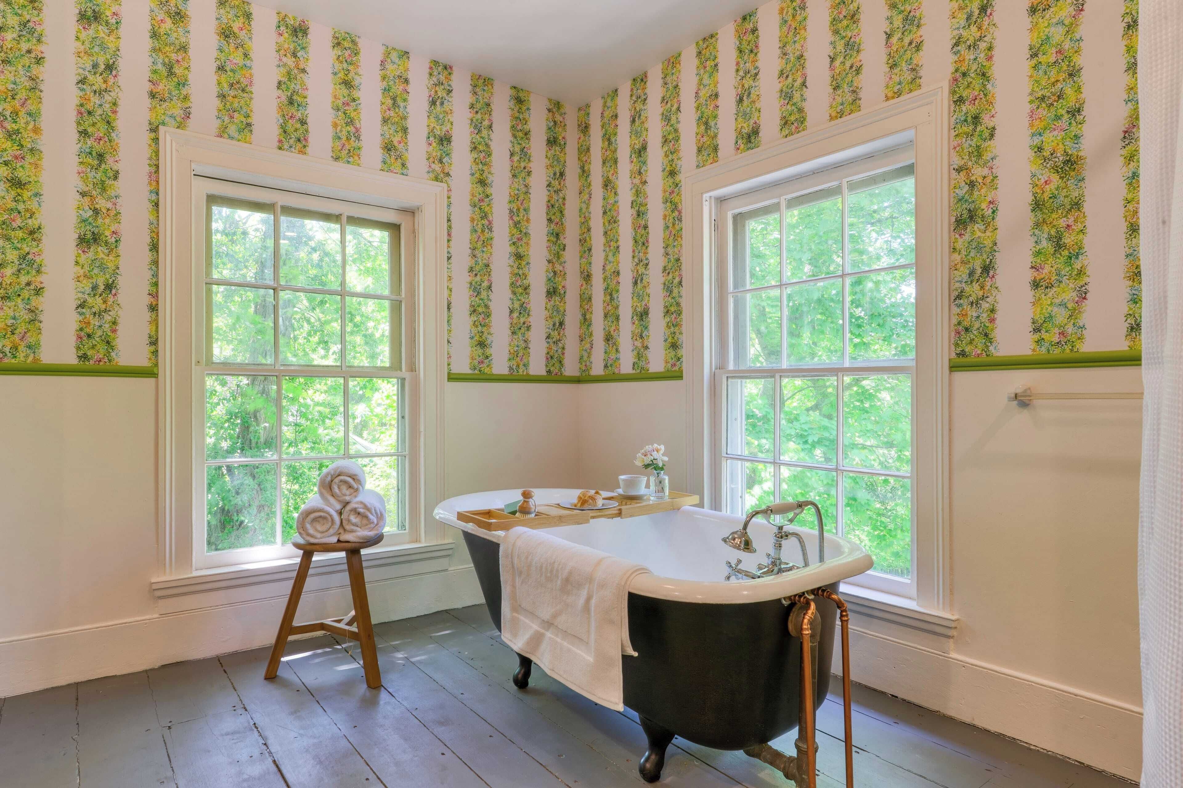 Kylpyhuone, jossa on jalkapäätyinen kylpyamme, kaksi ikkunaa, joissa on kukallinen tapetti, ja puinen jakkara, jonka päällä on pyyhkeitä.