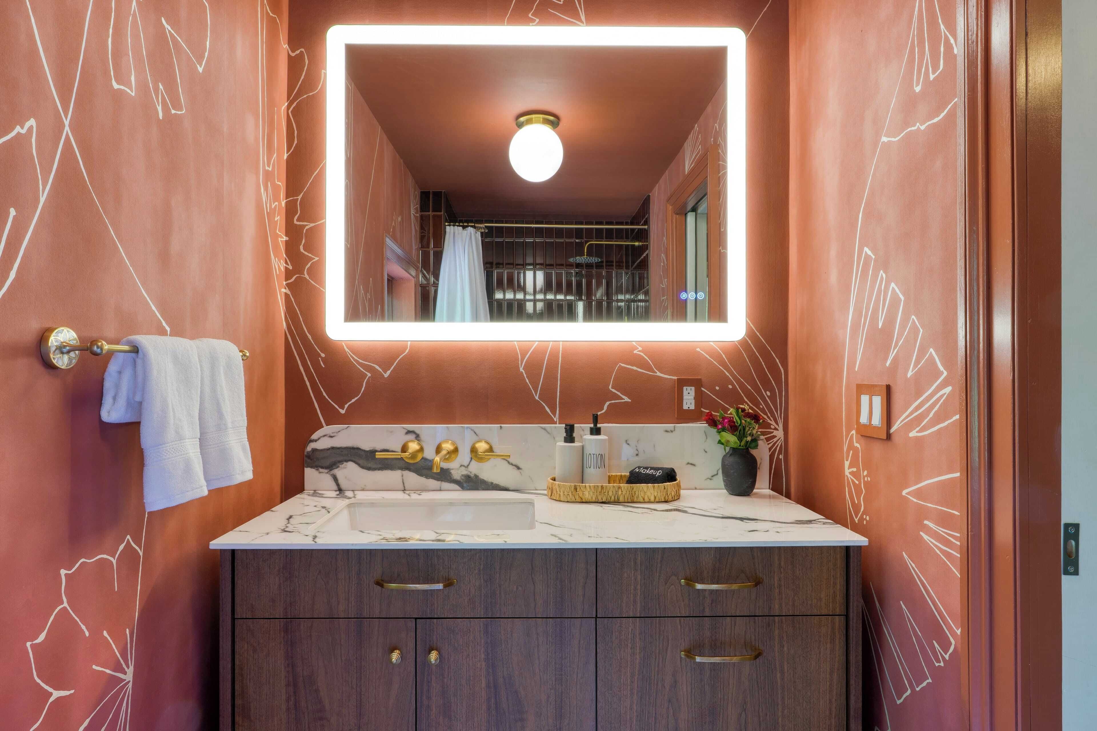 A bathroom with a marble sink, wooden cabinets, a large mirror with a light, and a towel rack.