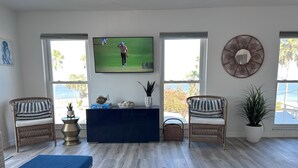Un salón con dos sillones de mimbre, un reposapiés azul, un mueble de entretenimiento azul oscuro y un televisor mostrando un partido deportivo.