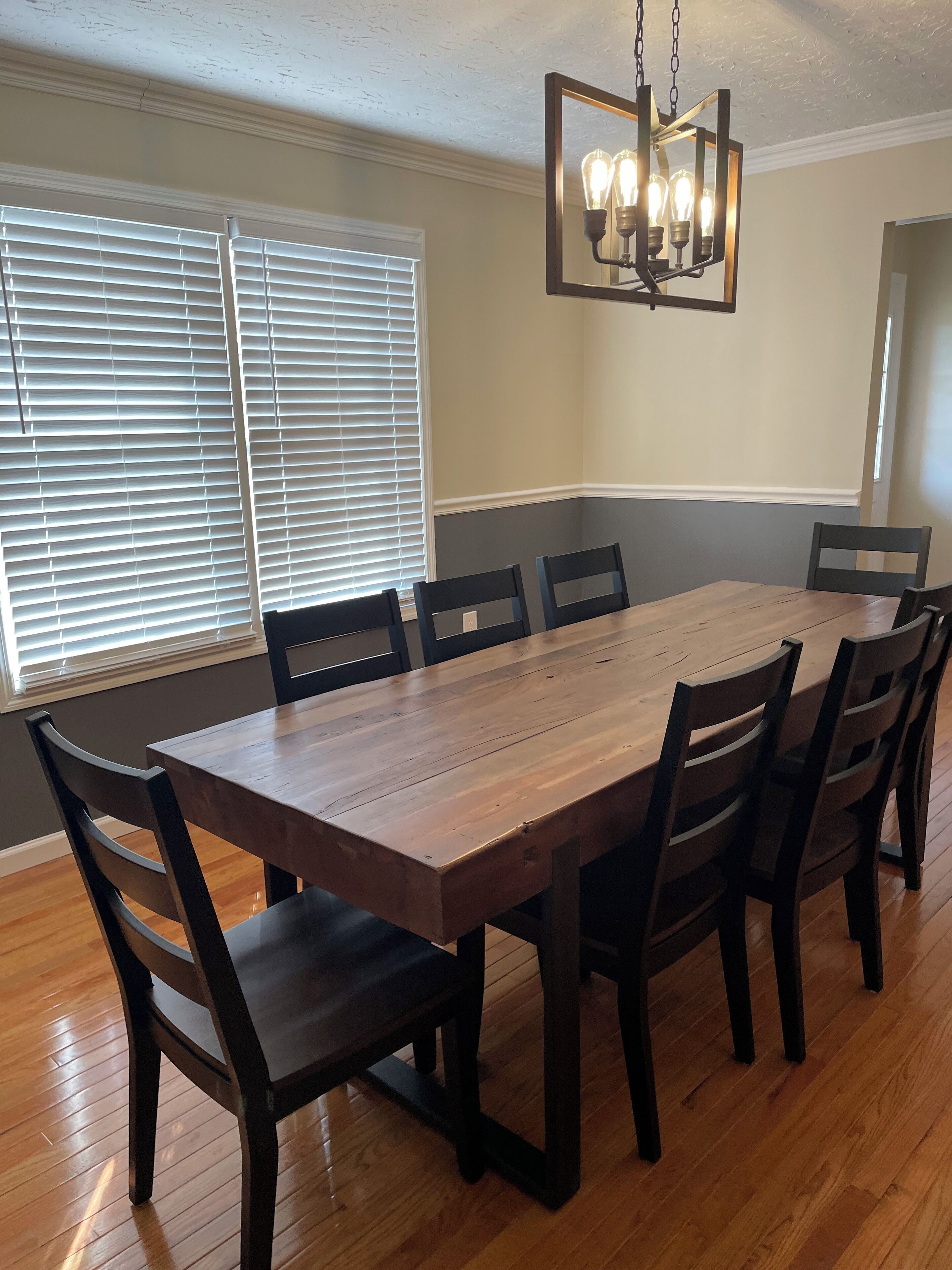 Une salle à manger avec une table et des chaises en bois, un lustre et une fenêtre avec des stores.