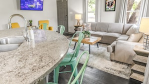 A modern living room with a kitchen area, including a granite countertop, green chairs, a beige sofa, and a television displaying a cityscape.