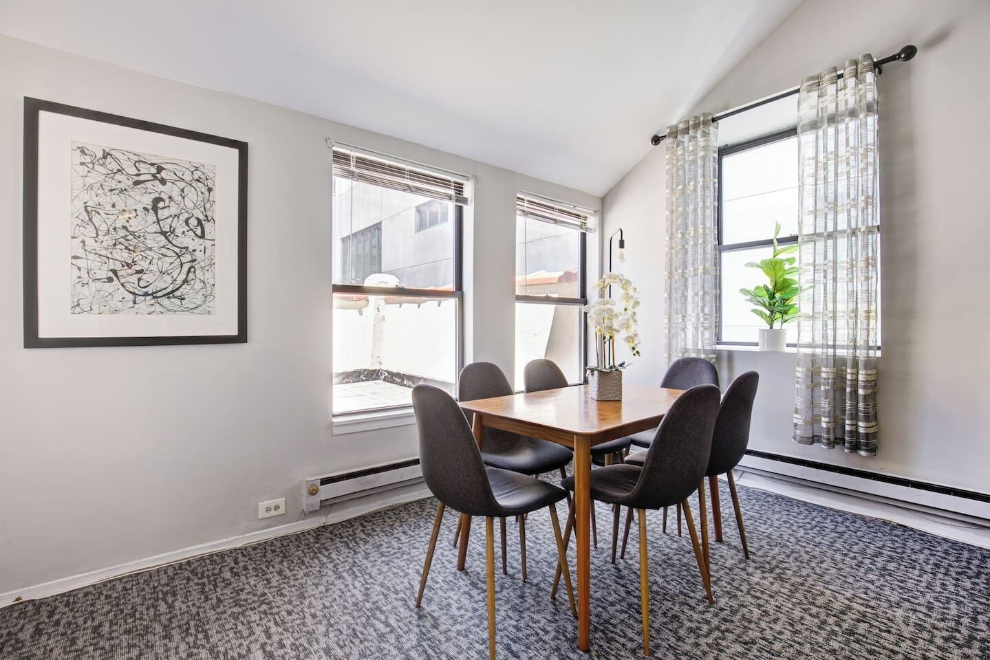 Een moderne eetruimte met een houten tafel en zwarte stoelen, grote ramen met zonwering en een lijst met abstracte kunst aan de muur.