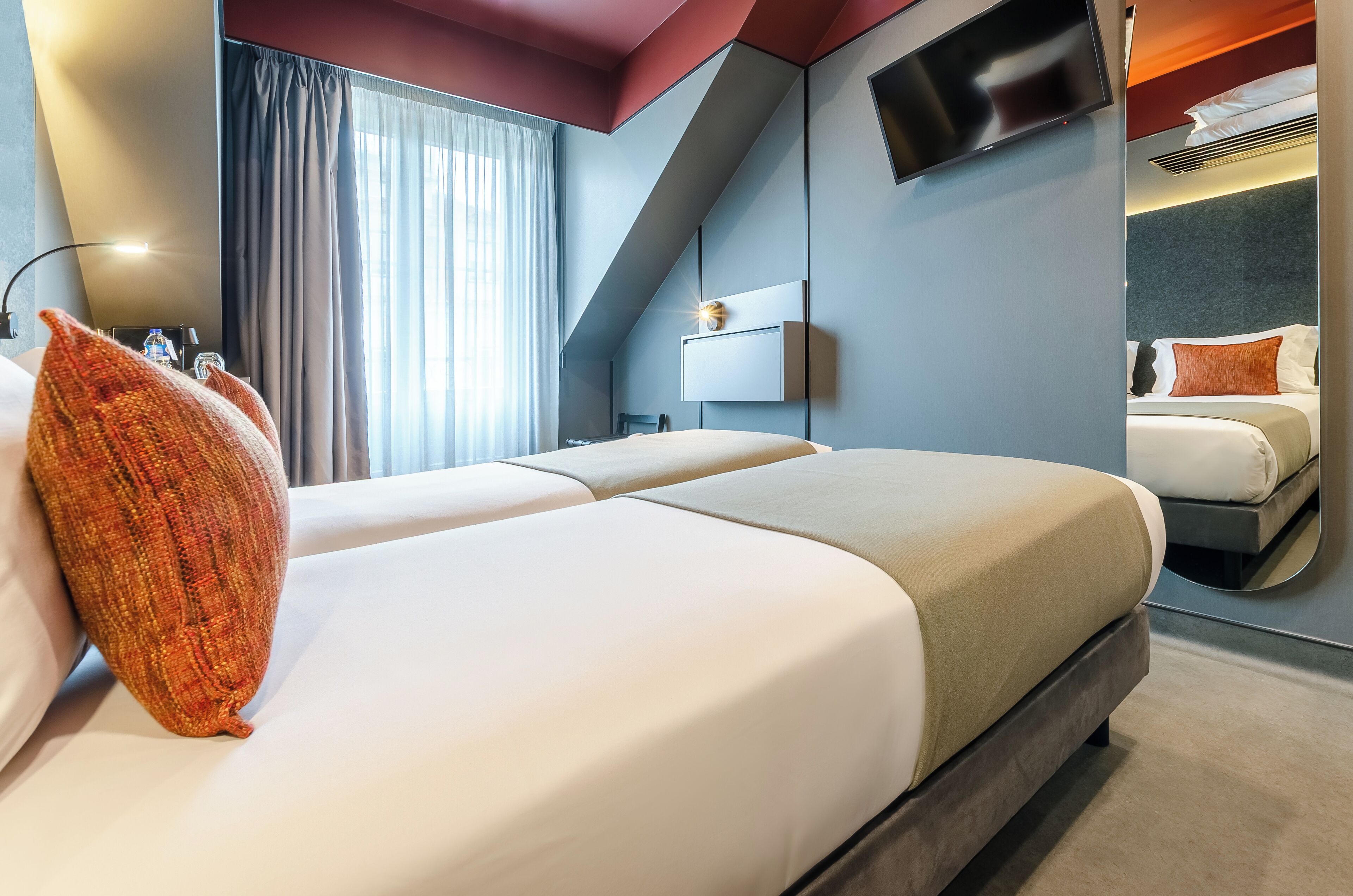 A modern hotel room with two beds, a large mirror, a flat-screen TV, and a large window with curtains.