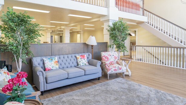 Ein geräumiger Wohnbereich mit einem grauen Sofa, einem blumigen Sessel und zwei Kübelpflanzen. Eine Treppe mit weißen Geländern führt auf eine Erschließung.