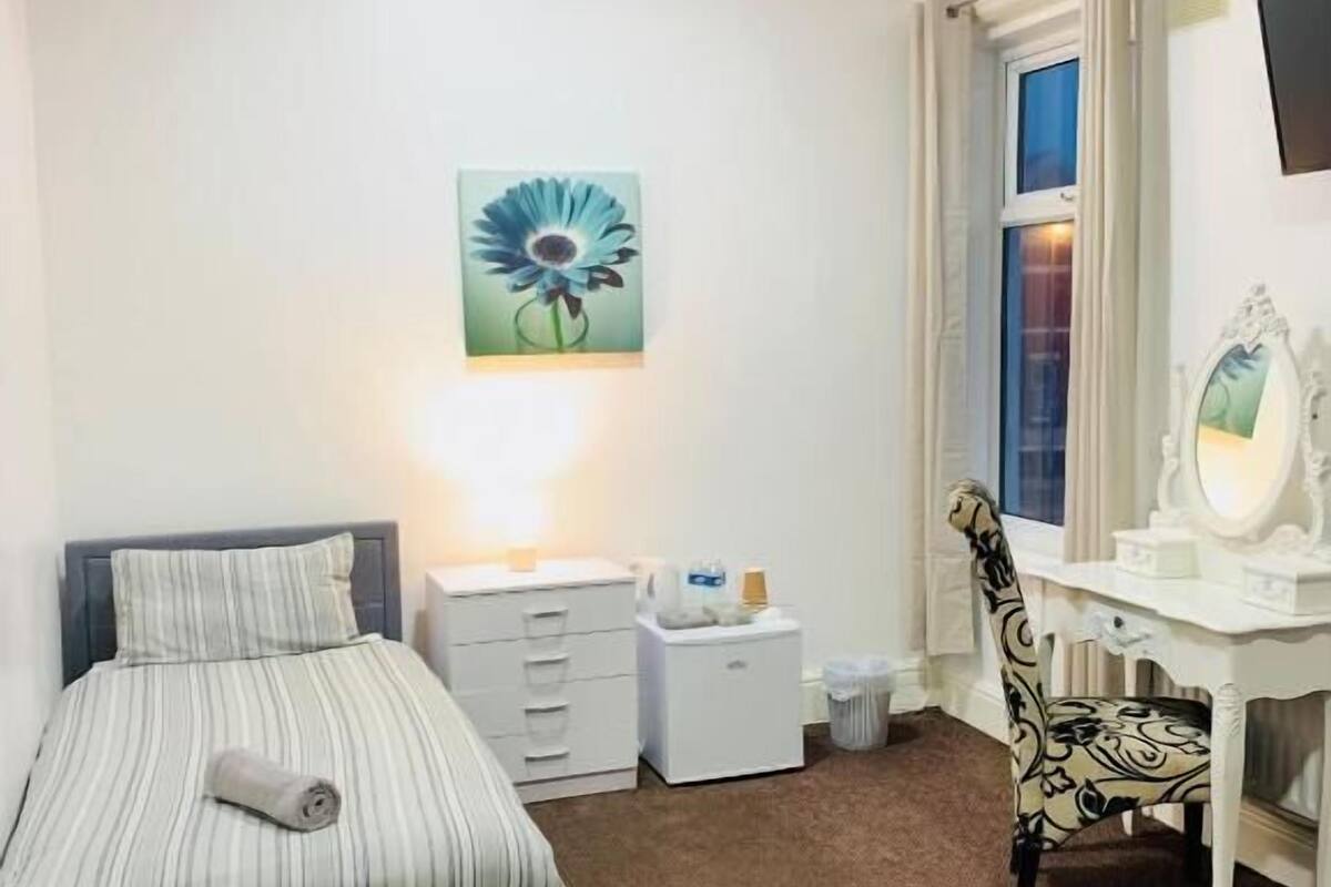 A bedroom with a bed, a desk, a chair, a nightstand, a lamp, a painting, and a window.