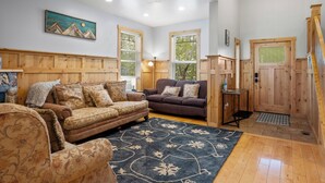 A cozy living room with a brown sofa, a patterned armchair, and a blue rug. There are wooden walls and a wooden door with a window. - Lakeside Haven, a cozy and relaxing home that sleeps 24 with pool/hottub access! (Garden City)