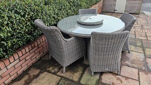 Una mesa redonda al aire libre con sillas de mimbre, una jardinera de ladrillo con follaje verde y una cerca de madera al fondo.