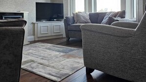 Una sala de estar con televisor de pantalla plana, dos sillones, un sofá, un tapete estampado y una araña.