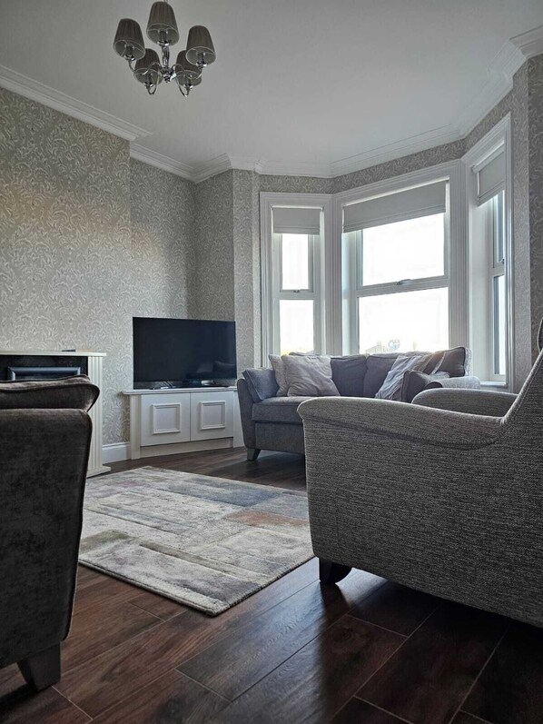 A living room with a flat-screen TV, two armchairs, a sofa, a patterned rug, and a chandelier.
