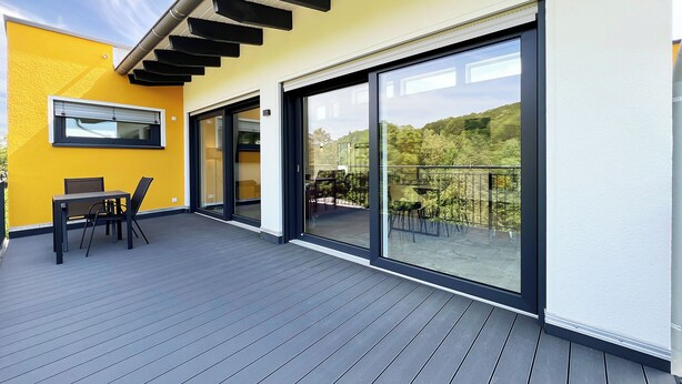 A modern outdoor patio with a yellow wall, a table with chairs, and a glass door leading to a balcony with a view of greenery. - Luxurious 4-room penthouse, roof terrace, parking space, e-charging station (Wachtberg)