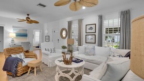 A spacious living room with a white sofa, a wicker chair, and a round coffee table. There is a ceiling fan, a window with curtains, and a painting on the wall. - 2 Side-by-Side Delray Beach Homes & Guest House - 1 blk to beach & Atlantic Ave! (Delray Beach)