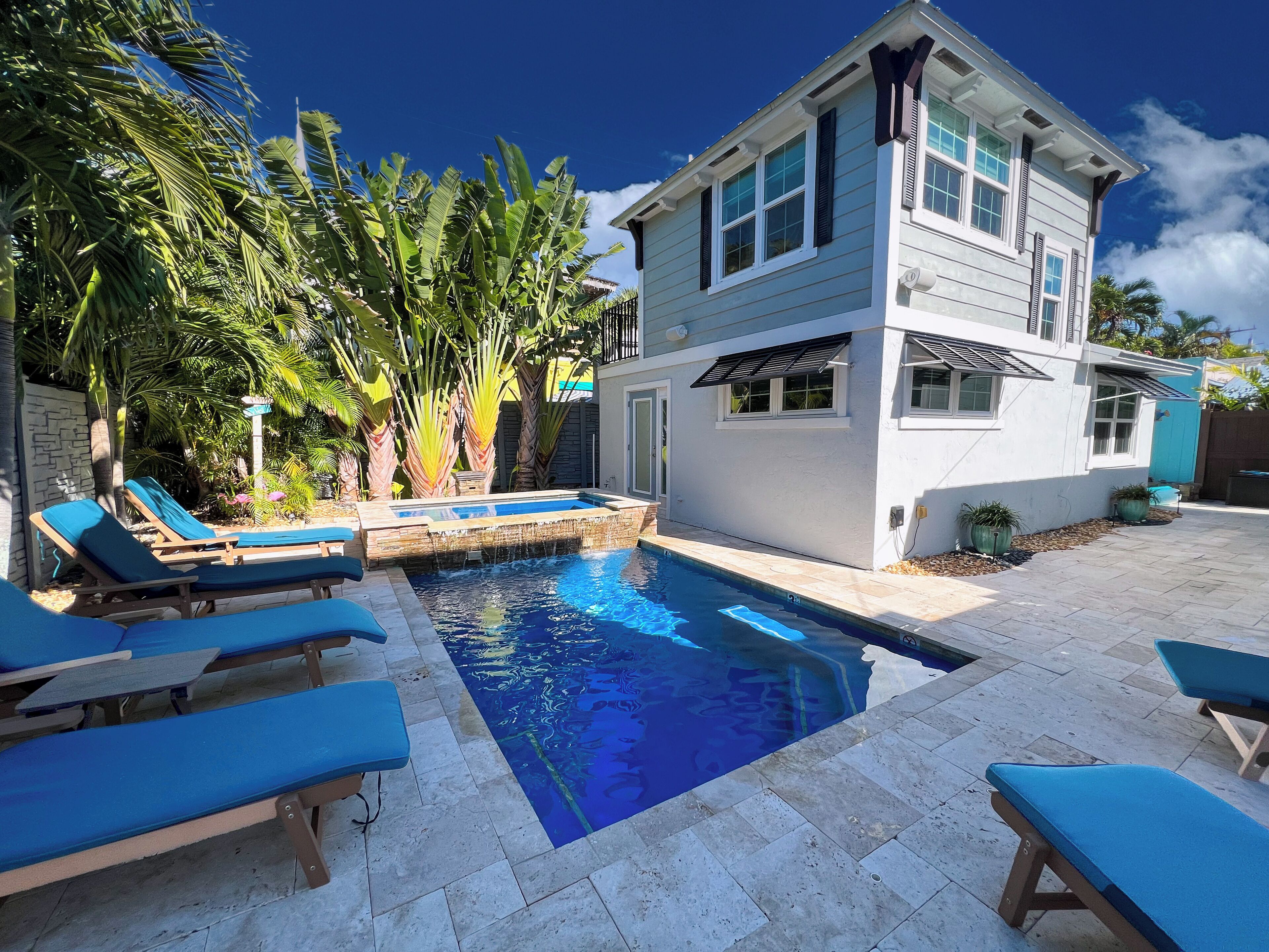 Ein zweistöckiges Haus mit Swimmingpool, blauen Liegestühlen und tropischen Pflanzen.