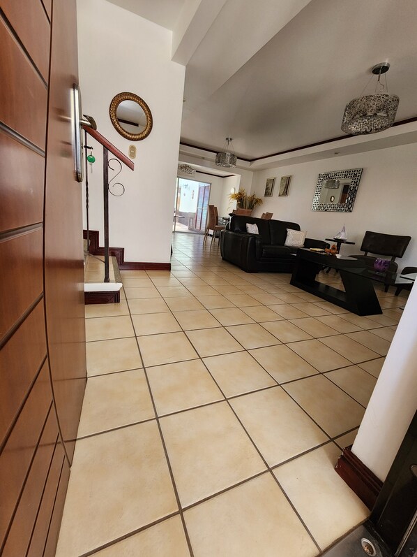 A spacious living area with a tiled floor, a black leather sofa, a dining table, and a staircase leading to another room.