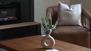 Un salon moderne avec une table basse en bois, un vase avec une plante, un canapé en cuir agrémenté d’un coussin et une cheminée en arrière-plan.