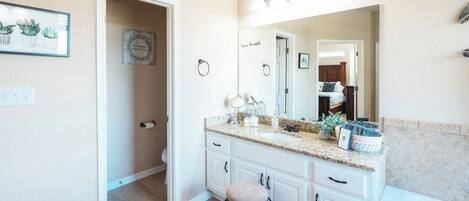 A bathroom with a vanity, sink, mirror, and a small stool. There is a shower area visible in the background.