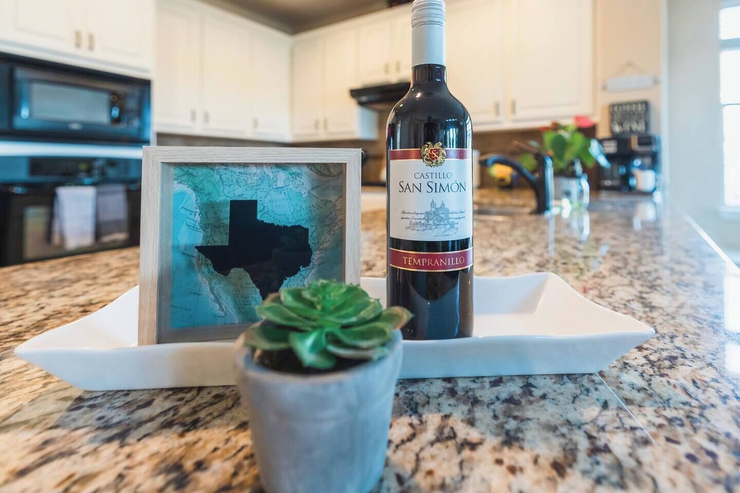 A bottle of Castillo San Simon Tempranillo wine on a white tray with a framed map of Texas and a succulent plant.