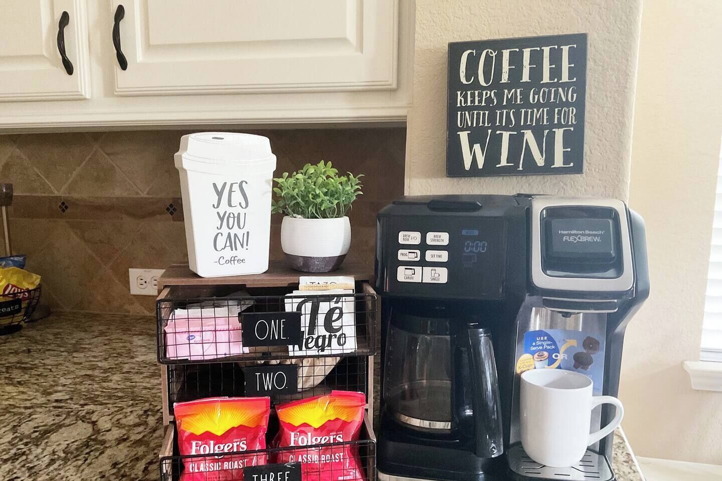 Una encimera de cocina con una cafetera, una taza y paquetes de café. Hay un cartel que dice 'El café me mantiene funcionando hasta que es hora de vino'.