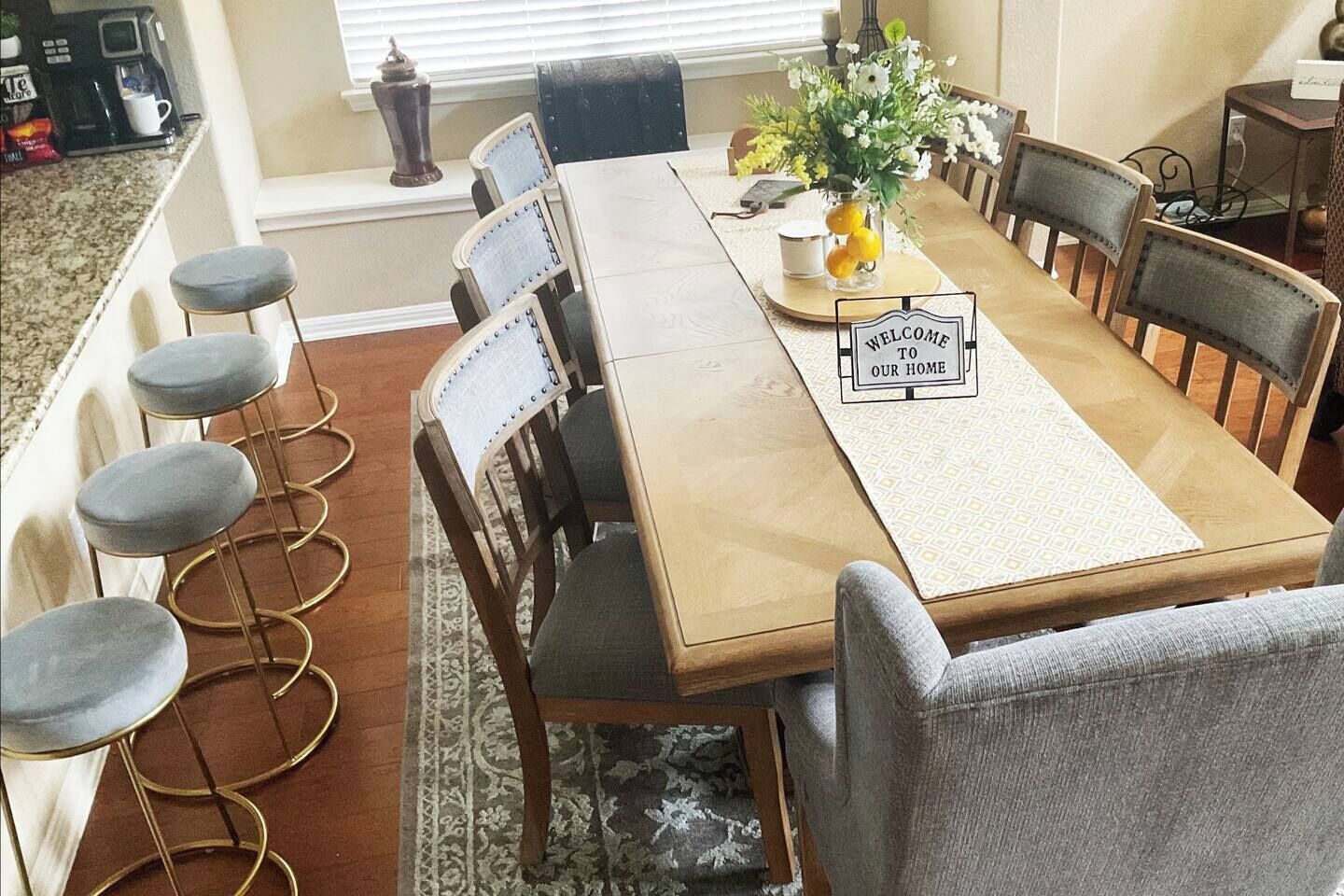 A dining area with a wooden table, four bar stools, and a 'WELCOME TO OUR HOME' placemat.