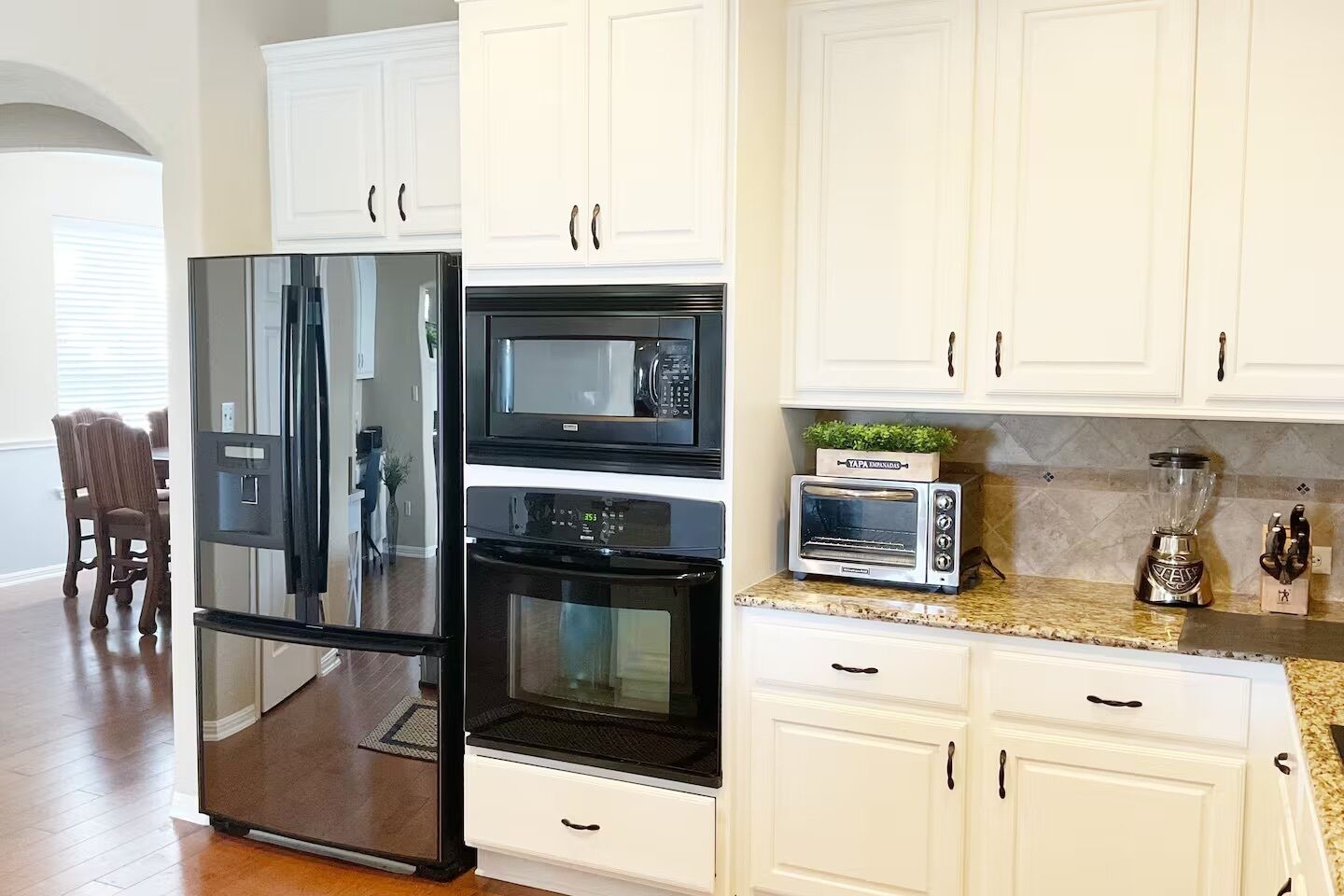 Una cocina con gabinetes blancos, un refrigerador negro, un microondas, un tostador y una licuadora.