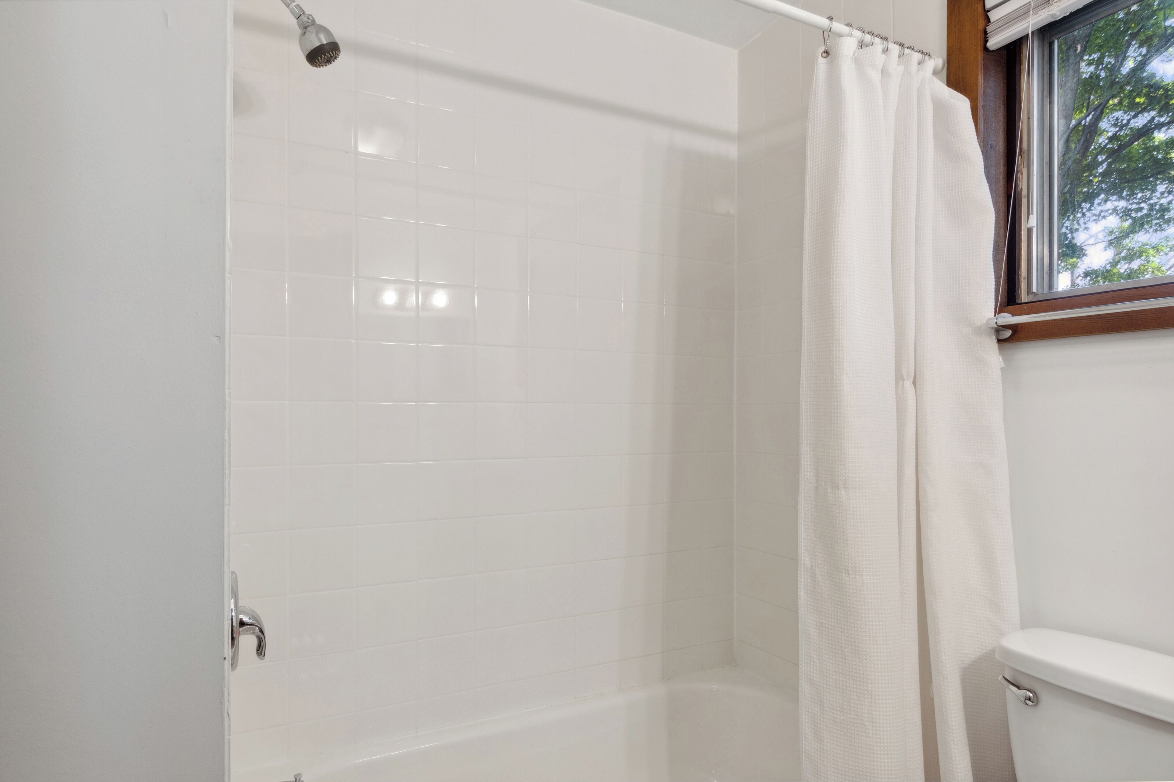 A bathroom with white tiled walls, a shower with a curtain, and a window with a view of trees.