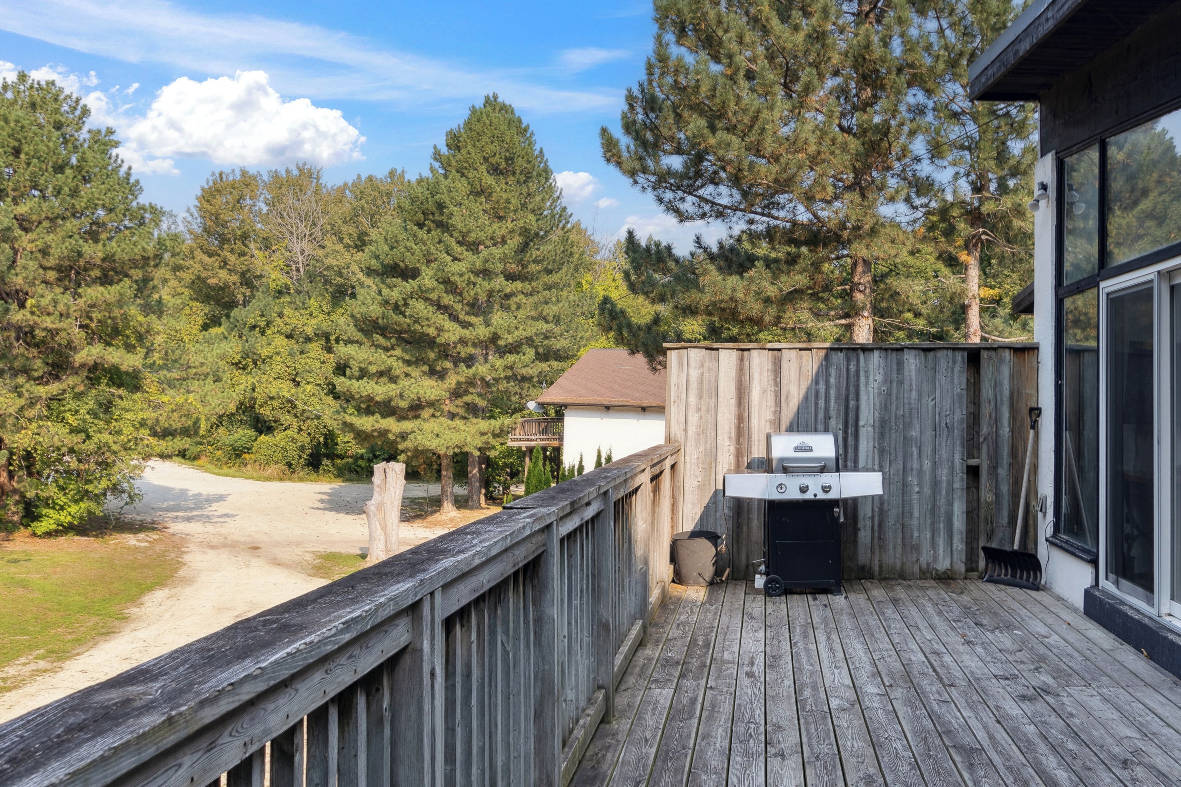 A wooden deck with a grill, overlooking a forested area and a dirt path.