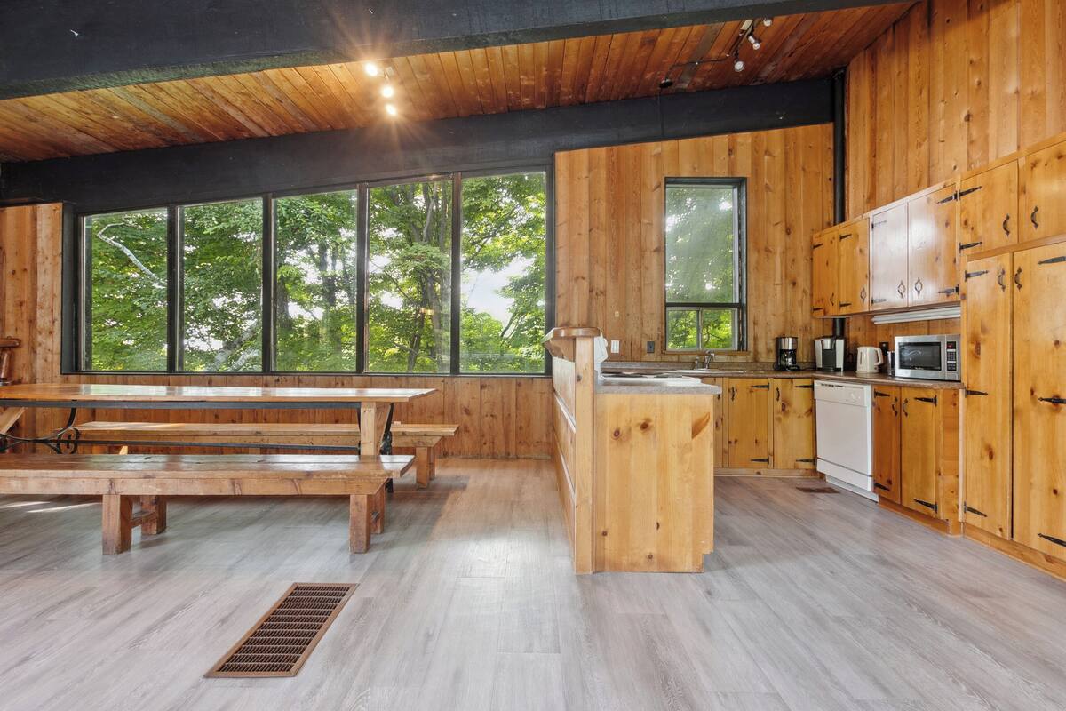 A modern kitchen with wooden cabinets, a dining area with a wooden table and benches, and large windows offering a view of trees.
