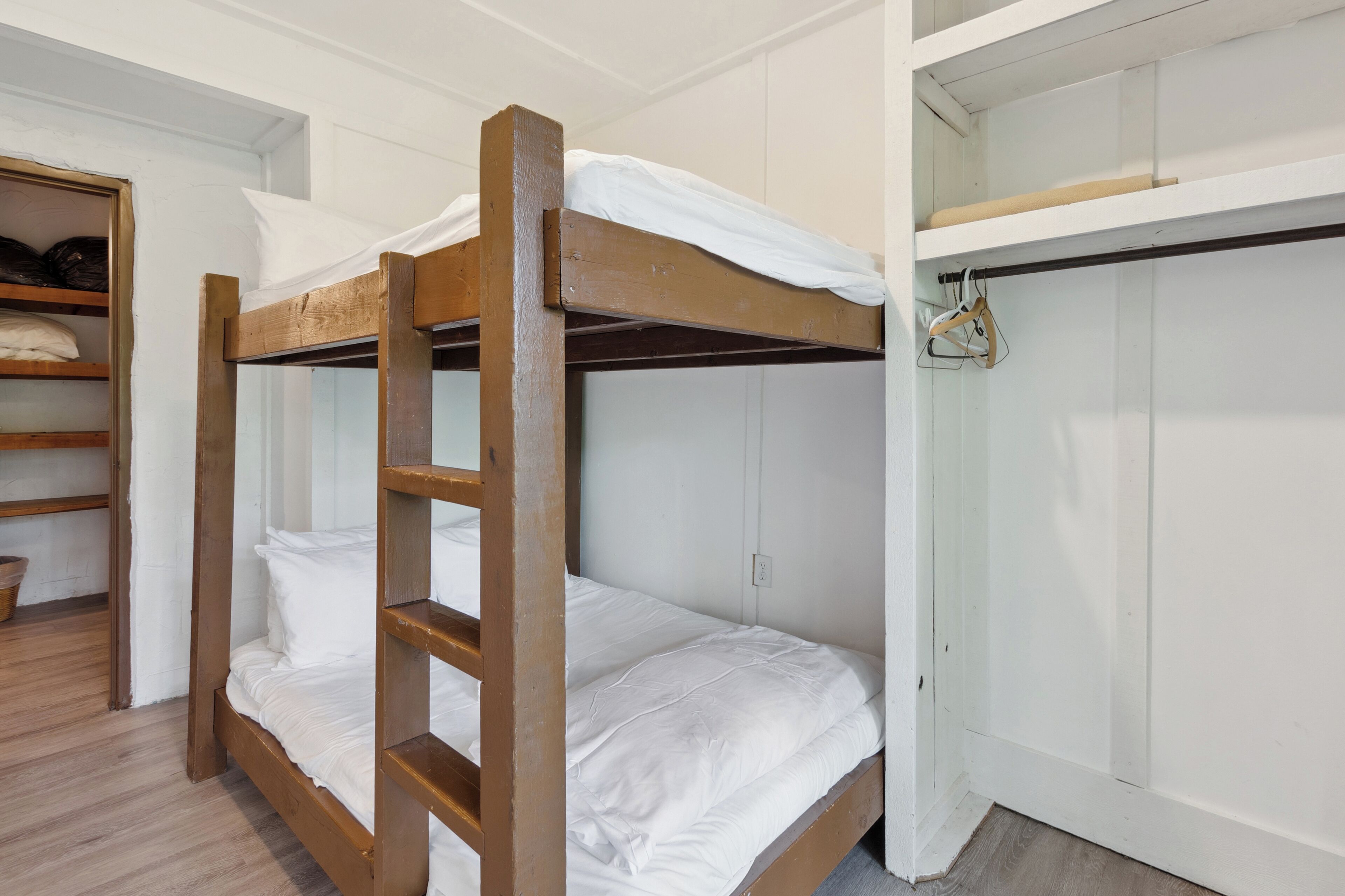 Uma cama beliche com roupa de cama branca, uma escada de madeira e um armário com espaço para pendurar.