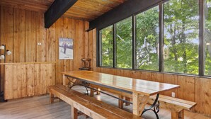Een houten interieur met een grote tafel en banken, een poster aan de muur, en een raam met uitzicht op bomen.