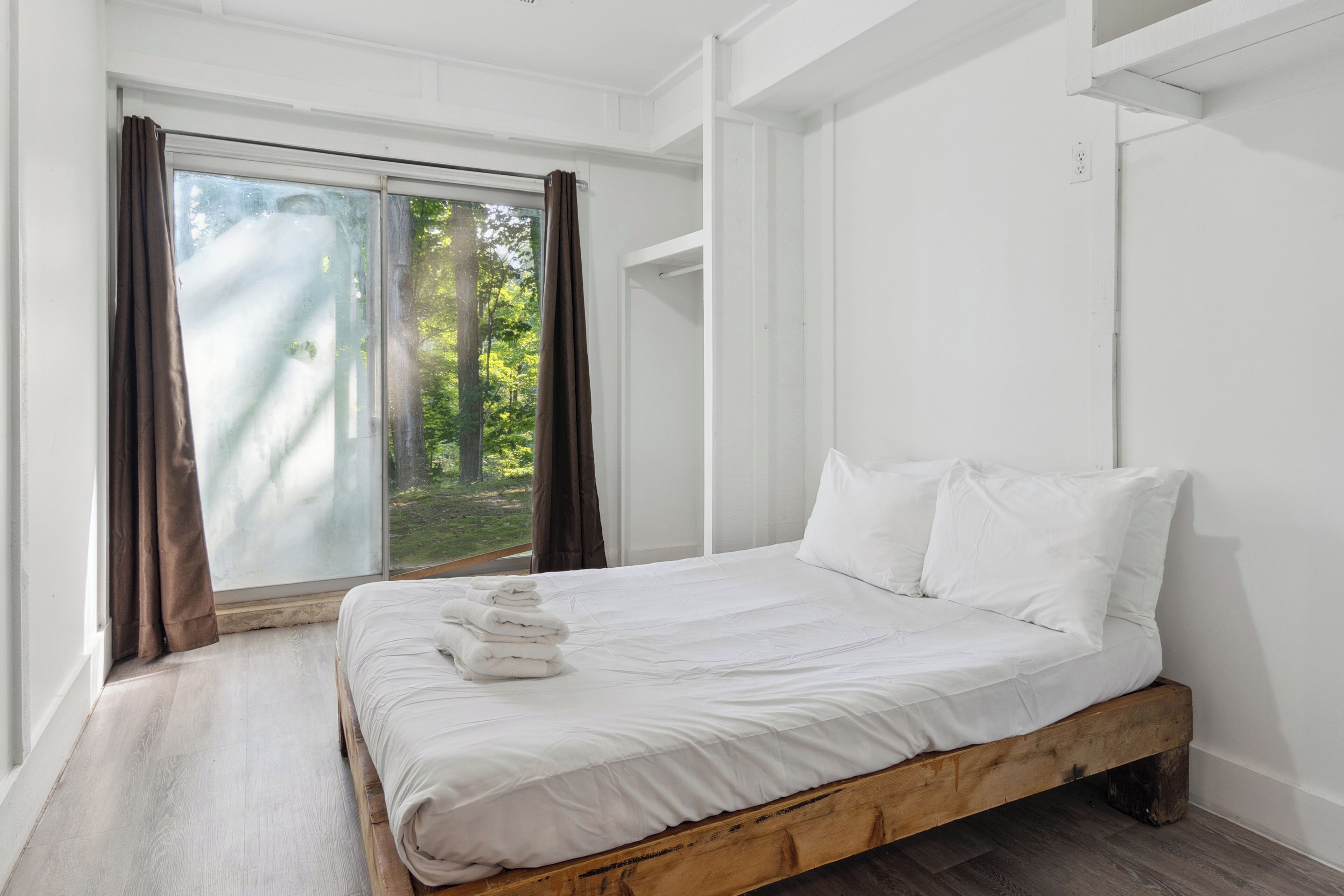A bedroom with a wooden bed frame, white bedding, and a view through a large window with curtains.