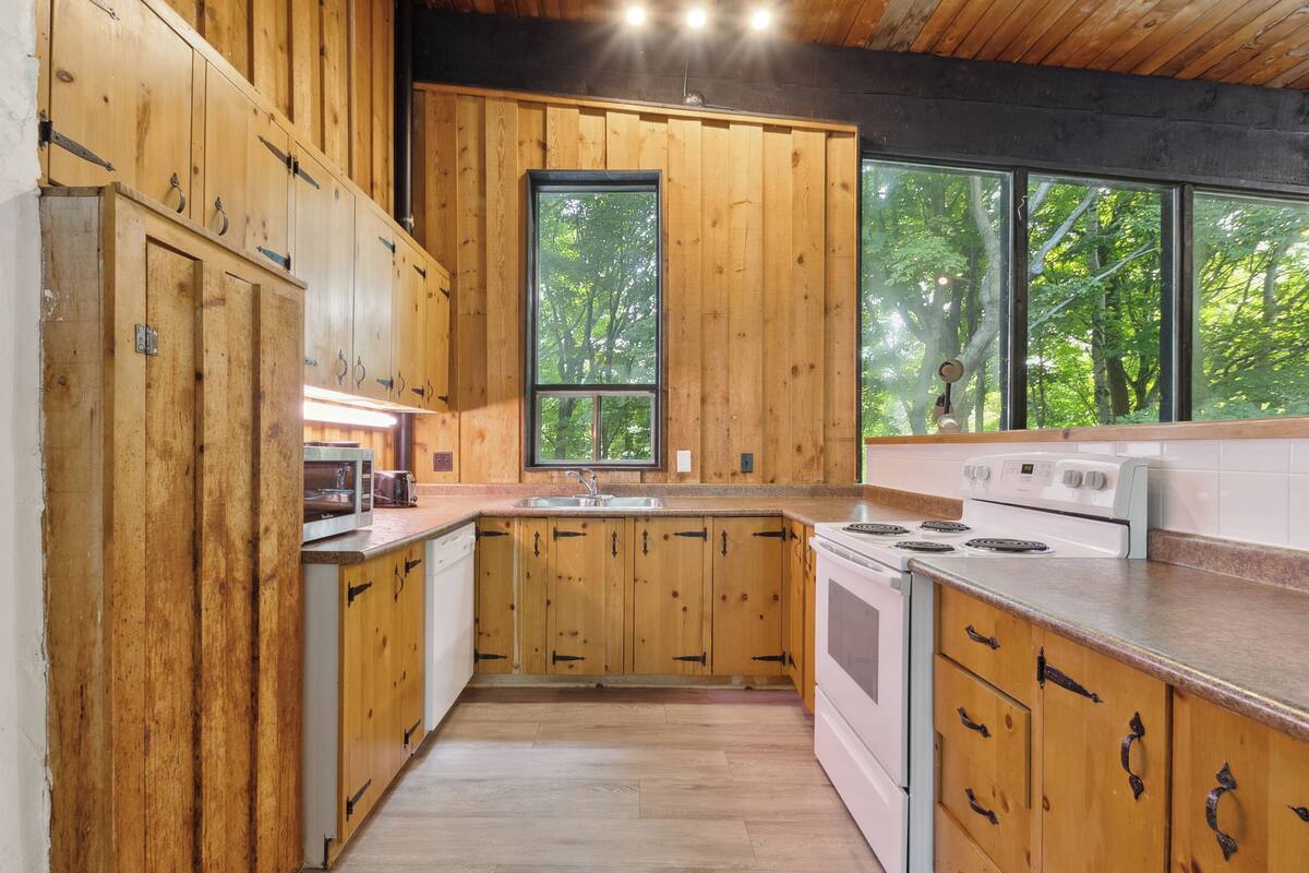 A kitchen with wooden cabinets, a white stove, and a large window overlooking a forest.