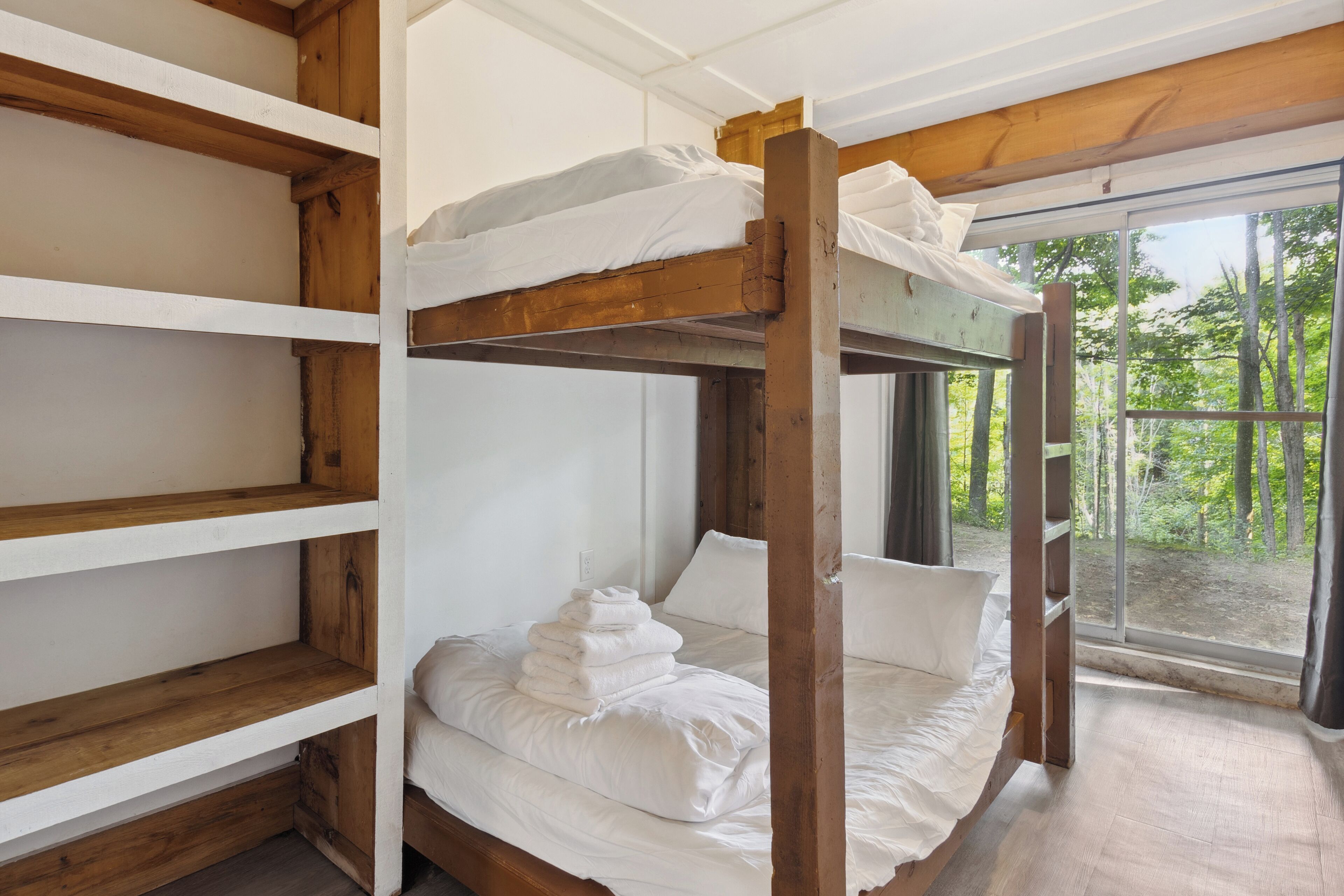 Ein Stockbett mit weißer Bettwäsche und einem Stapel Handtüchern auf der unteren Liege. An beiden Seiten des Bettes befinden sich Holzregale.