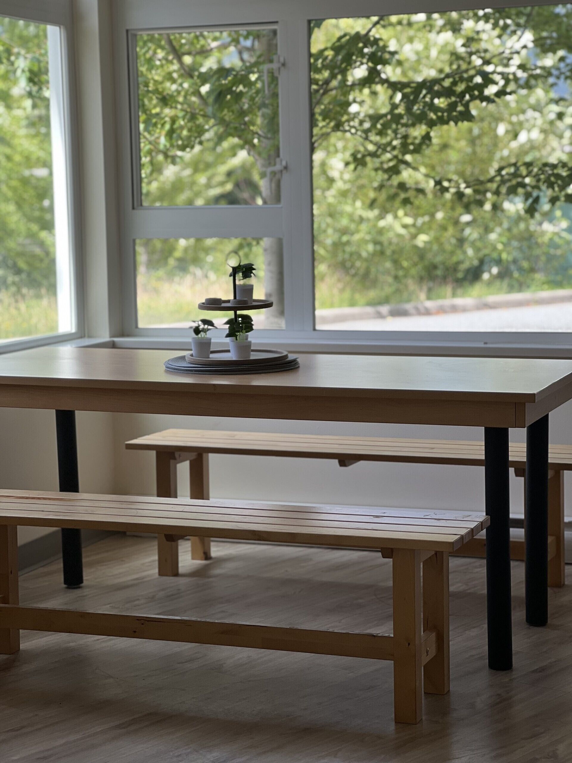 Una mesa de madera con bancos, un estante escalonado para plantas y vistas de vegetación a través de una ventana.