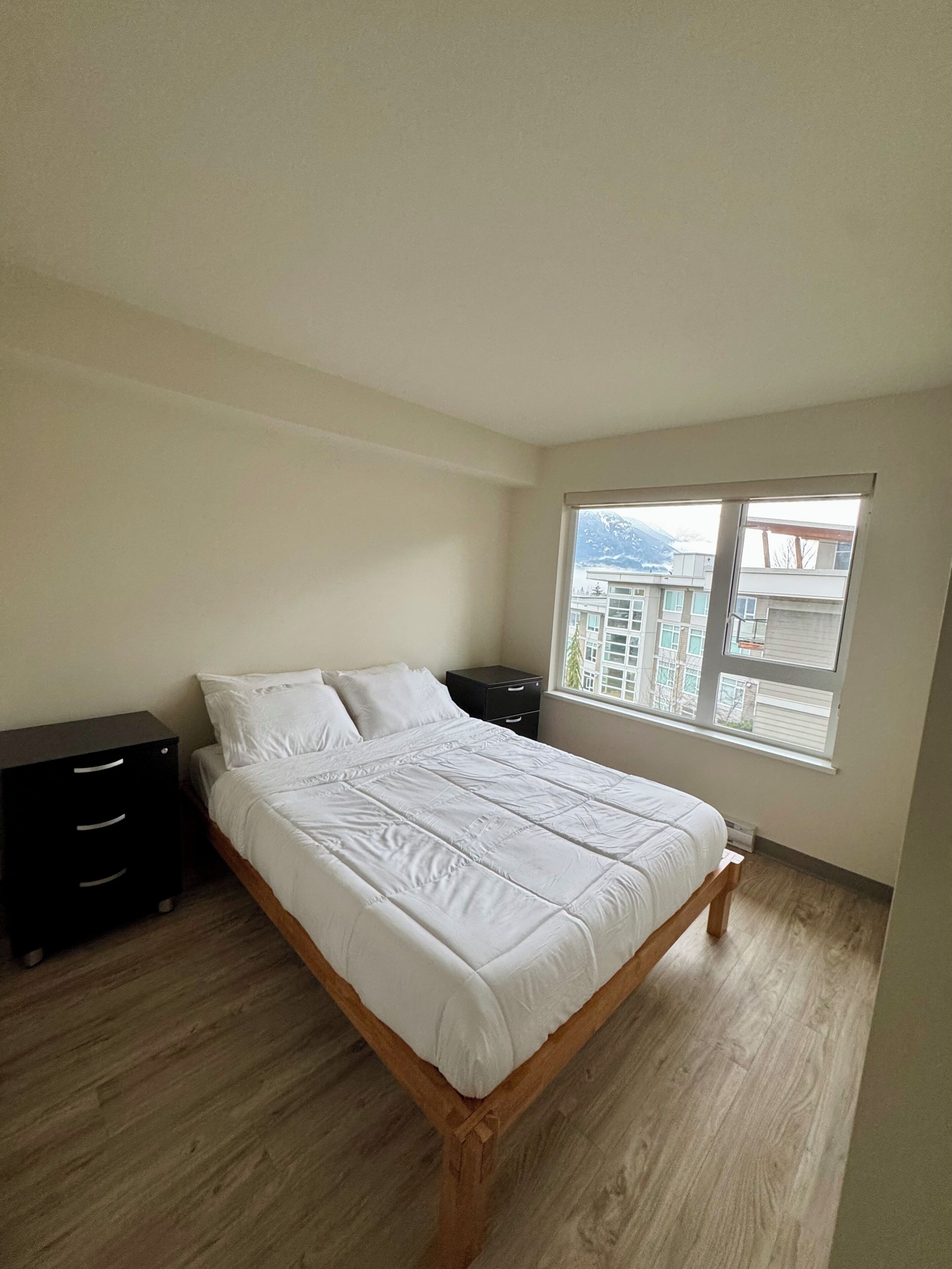 Un dormitorio con una cama de madera, una mesita de noche, una ventana con vistas a edificios y suelo de madera.