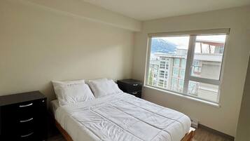 Un dormitorio con una cama de madera, una mesita de noche, una ventana con vistas a edificios y suelo de madera.
