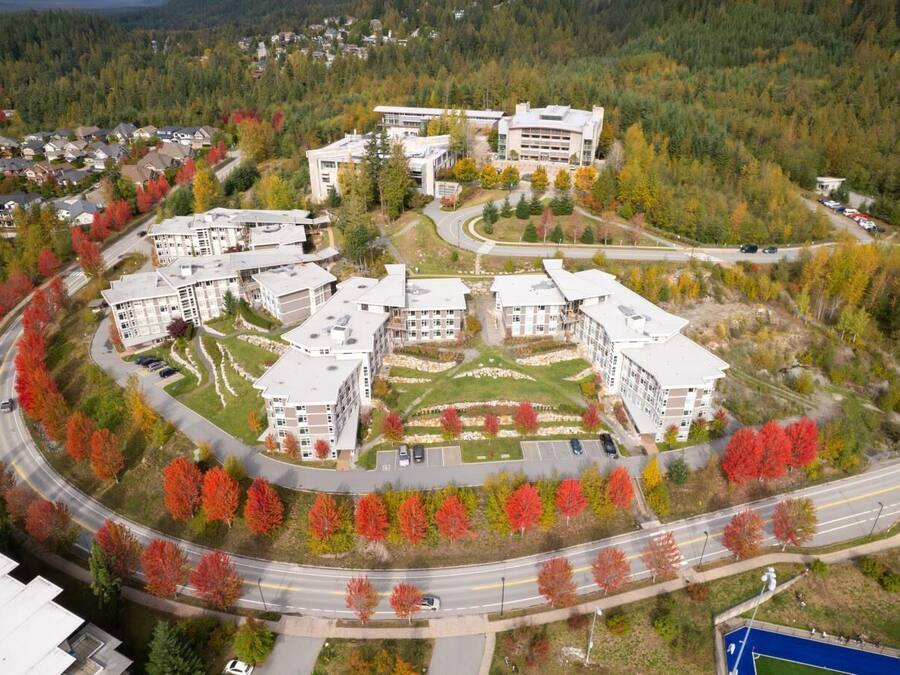Vista aérea de um campus com vários edifícios, ruas e área de estacionamento, tudo cercado por árvores com folhagem outonal.