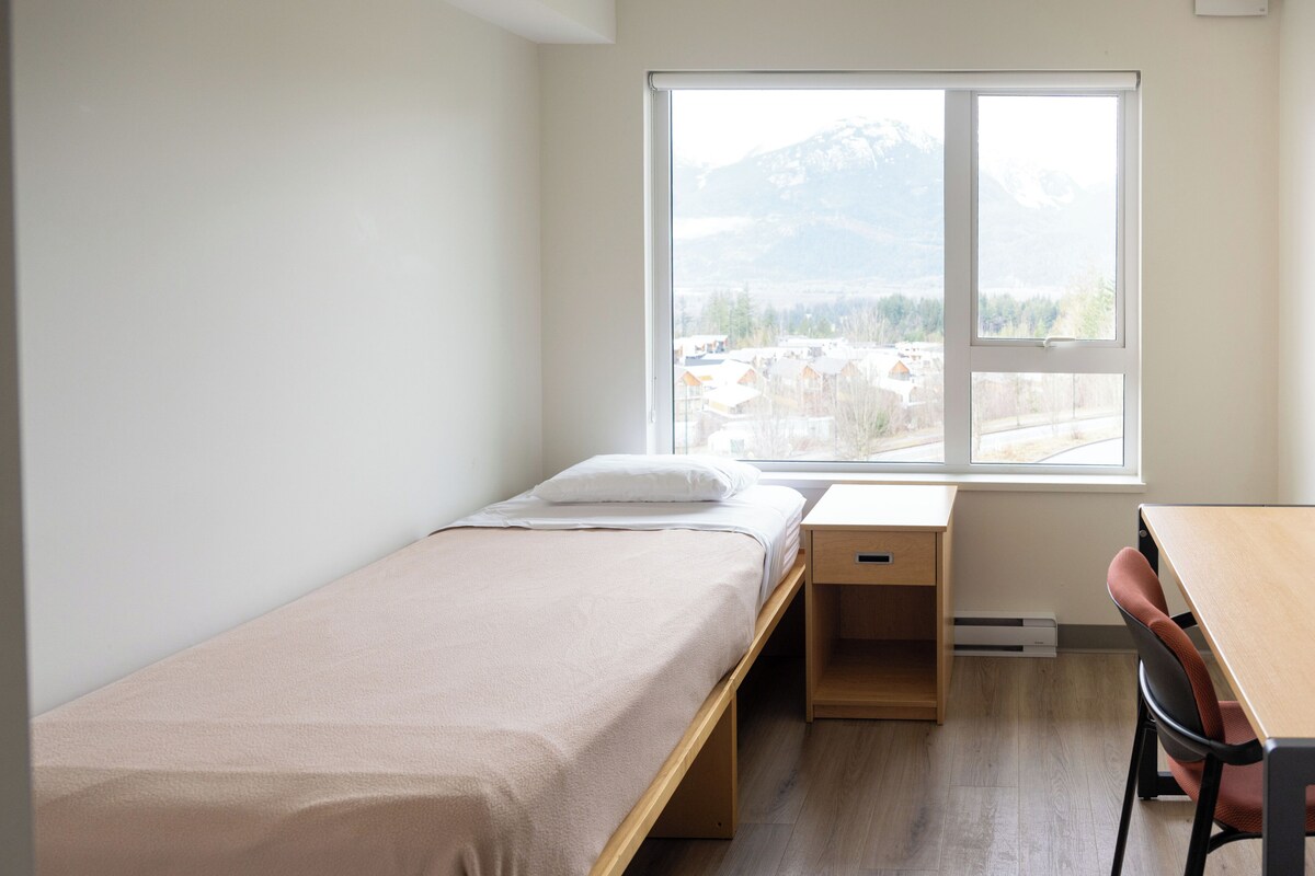 A small, single-bed room with a wooden desk and chair, a window with a view of a mountain and buildings, and a white wall.