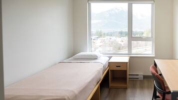 Una habitación pequeña con cama individual, un escritorio y una silla de madera, una ventana con vistas a una montaña y edificios, y una pared blanca.