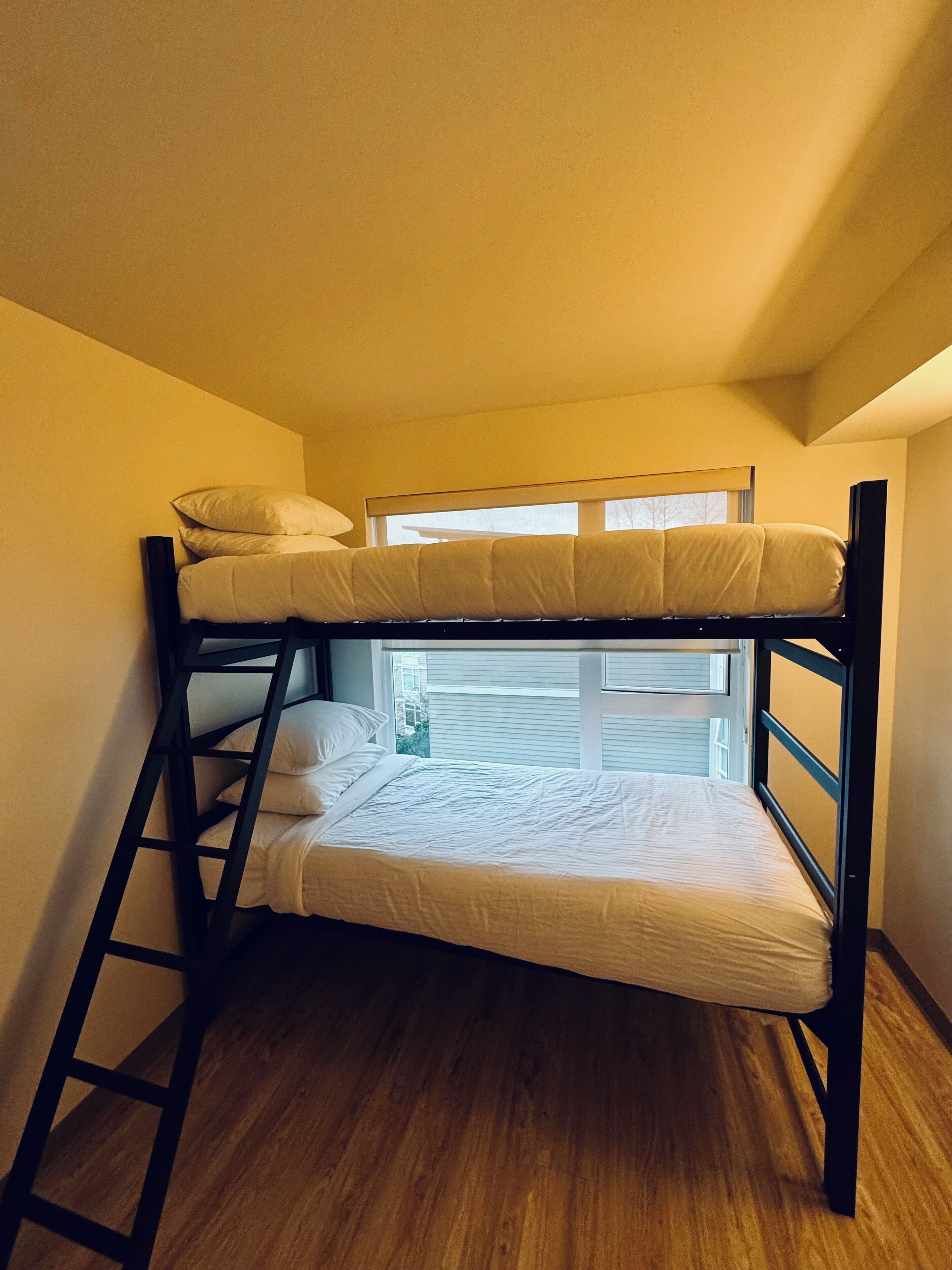 Uma cama de beliche com escada, janela com persianas e vista para o exterior.