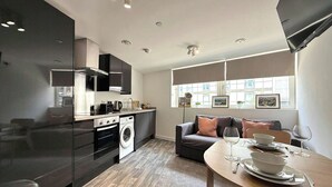 A modern kitchen with a built-in oven, washing machine, and black cabinets. There is a dining area with a table set for two, and a sofa in the background. - City Centre 1Bed Flat (Sheffield)