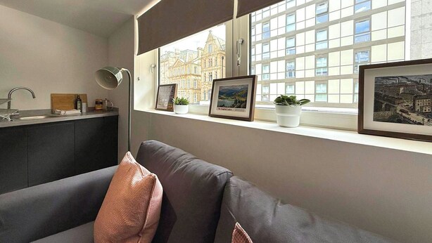 A modern living room with a grey sofa, a window with blinds, a kitchen area, and framed pictures on the windowsill. - City Centre 1Bed Flat (Sheffield)