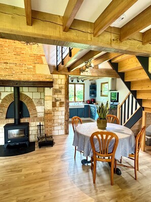 A rustic interior with exposed wooden beams, a stone fireplace, a dining area with a round table, and a kitchen in the background.