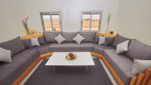 A modern living room with a grey sectional sofa, a white coffee table, and a view of a desert landscape through large windows. - Family Stay Ouarzazate (Ouarzazate)
