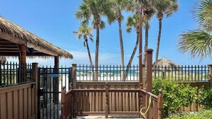 A gated entrance with a wooden fence, a thatched roof structure, and a view of a beach with palm trees and umbrellas.