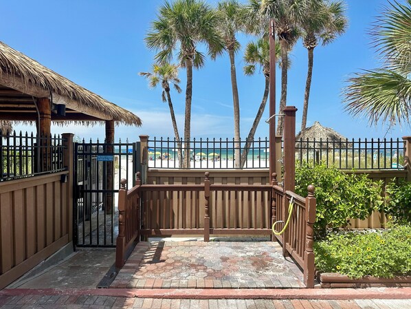 A gated entrance with a wooden fence, a thatched roof structure, and a view of a beach with palm trees and umbrellas.