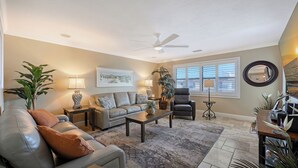 A living room with a sofa, armchairs, a coffee table, and a floor lamp. There is a ceiling fan, a painting on the wall, and a window with blinds.