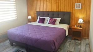 Een slaapkamer met een groot bed, houten paneelwanden, een raam met zonwering en een nachtkastje met een lamp.
