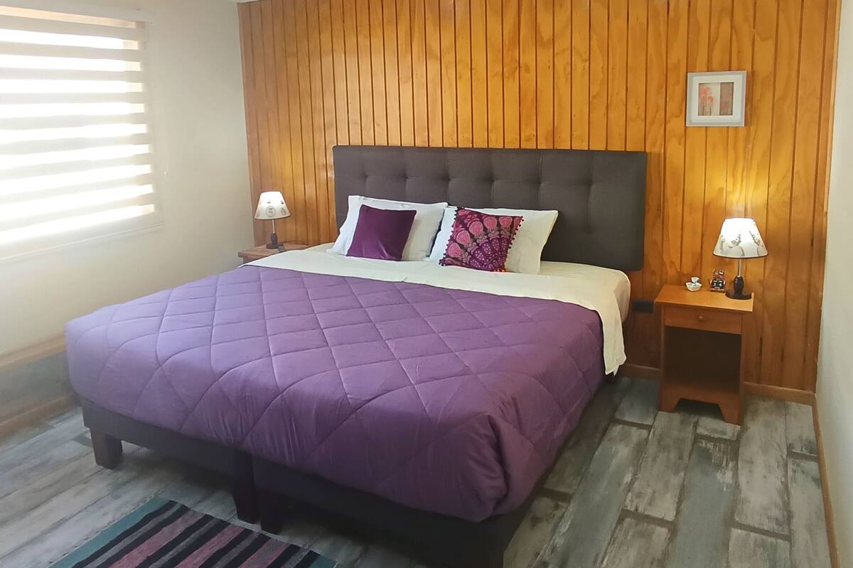Une chambre à coucher avec un grand lit, des murs recouverts de panneaux de bois, une fenêtre avec des stores et une table de chevet avec une lampe.