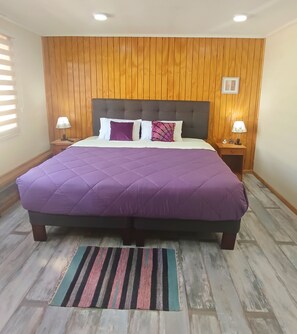 Een groot bed met een paars sprei, twee nachtkastjes met lampen, houten paneelwanden en een gestreept vloerkleed op een houten vloer.